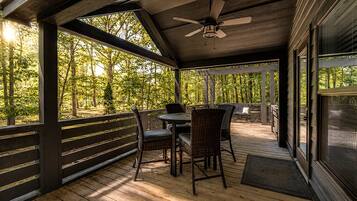 A covered outdoor area with a table and chairs, a ceiling fan, and a view of a wooded area.