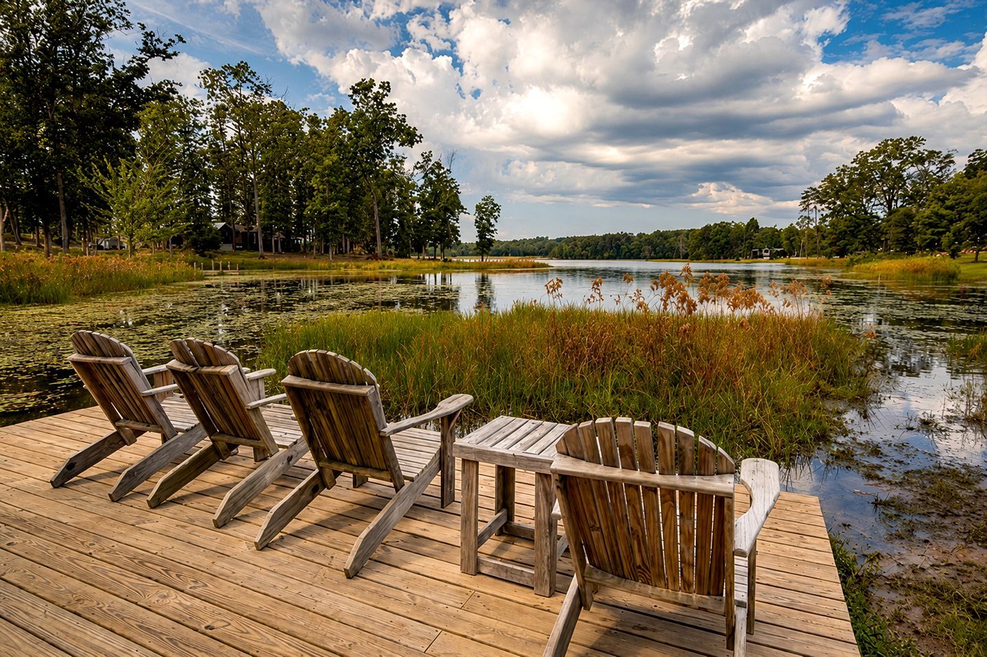 Un'area prendisole in legno con quattro poltrone Adirondack e un tavolo, affacciata su un lago tranquillo circondato da alberi.
