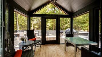 A covered outdoor seating area with a table and chairs, a view of a deck with a barbecue grill, and a wooden floor.