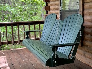 Una silla Adirondack verde sobre una terraza de madera con barandilla y una cabaña de troncos al fondo.