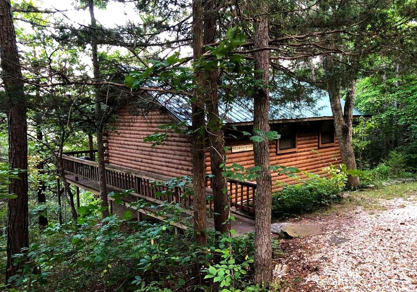 A log cabin with a wooden deck and a metal railing, surrounded by dense forest.