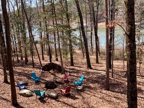 A forested area with several Adirondack chairs arranged in a circle, a pile of firewood, and a body of water in the background. - Waterfront Cabin Ideal for Families and Outdoor Adventures in Arkansas (Fairfield Bay)