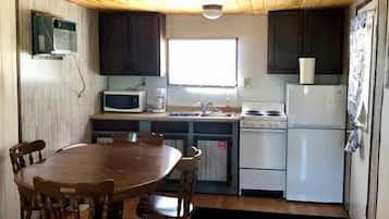 A compact kitchen with a dining table, wooden chairs, a microwave, a refrigerator, and a window providing natural light.
