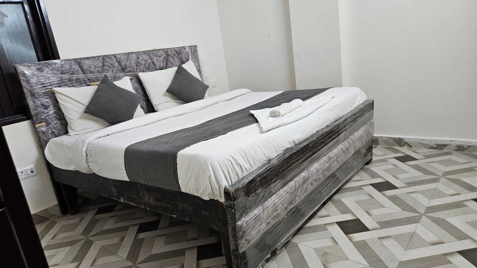 A bed with a wooden frame, white bedding, and grey pillows in a room with patterned floor tiles.