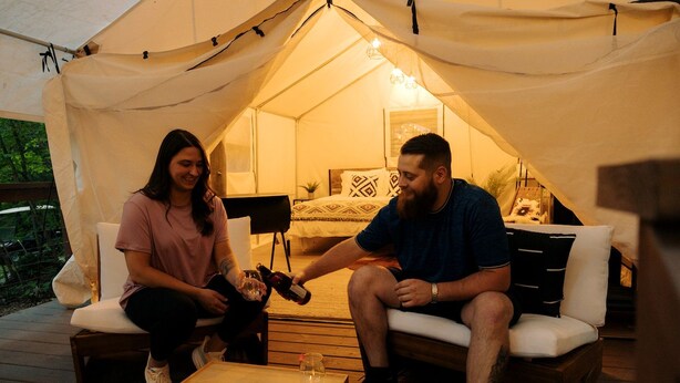 Alojamiento con carpa, terraza de madera, una pequeña mesa con velas y una cama con ropa de cama estampada.