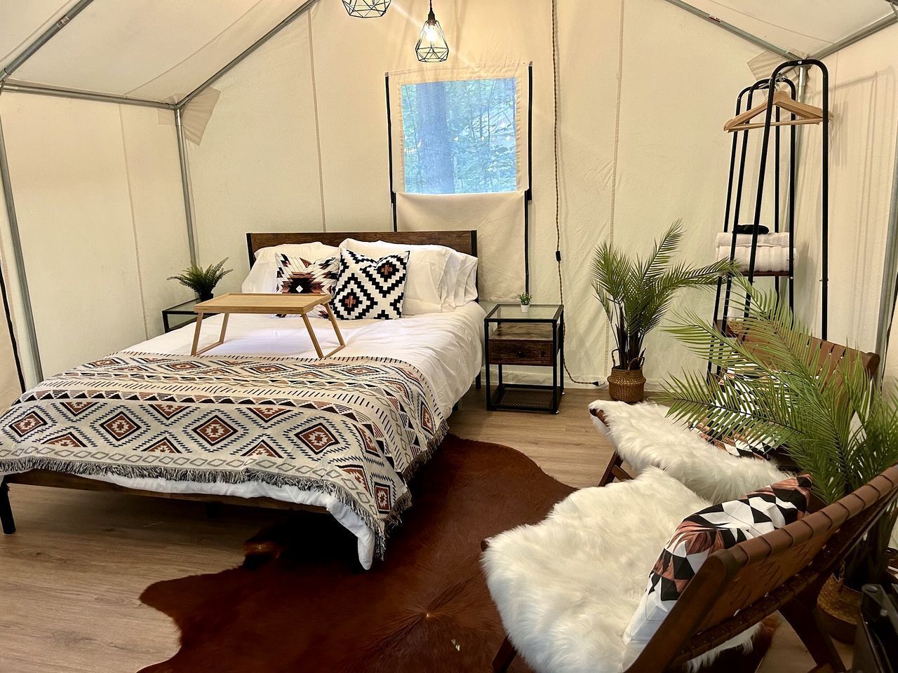 A tent-style room with a bed, a hanging light fixture, a small table, a chair, and a window with a view of trees.