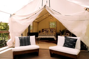 A tented accommodation with a bed, two cushioned seating areas, and a wooden deck.