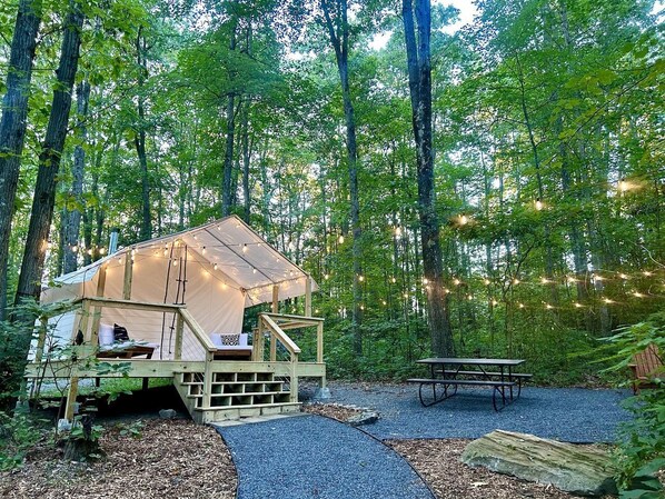 A forested campsite with a tent, wooden deck, picnic table, and string lights.