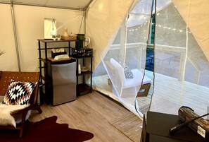 A tented room with a bed, a small table with a lamp, a refrigerator, and a chair with a cushion.