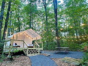 A forested campsite with a tent, wooden deck, picnic table, and string lights.