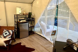 A tented room with a bed, a small table with a lamp, a refrigerator, and a chair with a cushion.