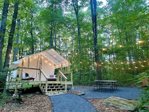 A forested campsite with a tent, wooden deck, picnic table, and string lights.