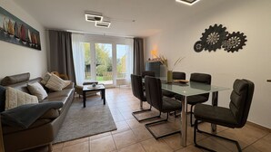 A modern living room with a sofa, a glass dining table, and chairs. There is a painting on the wall and a window with curtains. - Vacation home "MeerZeit" - Holiday home over 3 floors, 65 m² living space with terrace (Dahme)