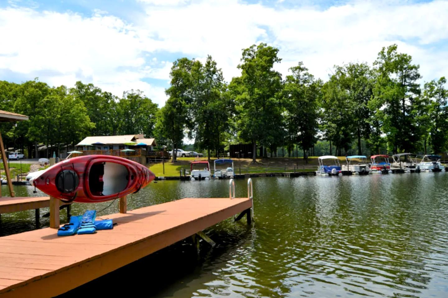 A dock with a kayak and paddleboards, a row of boats, and a park with trees and a building in the background.