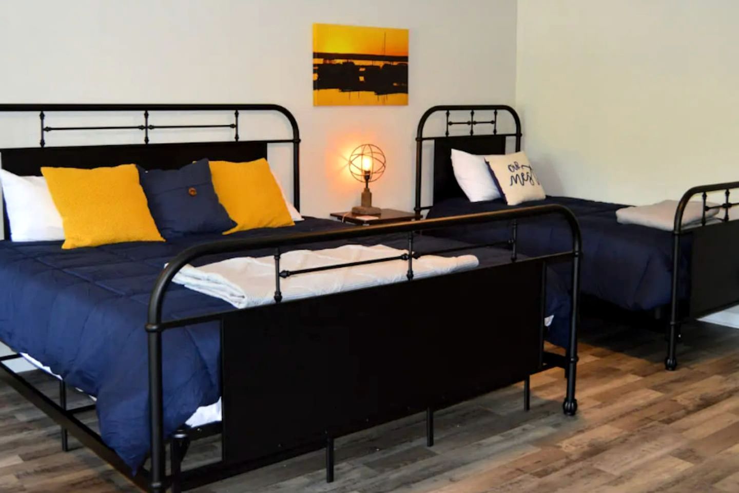 A double bed with a black metal frame, two pillows, a bedside table with a lamp, and a wall art piece.