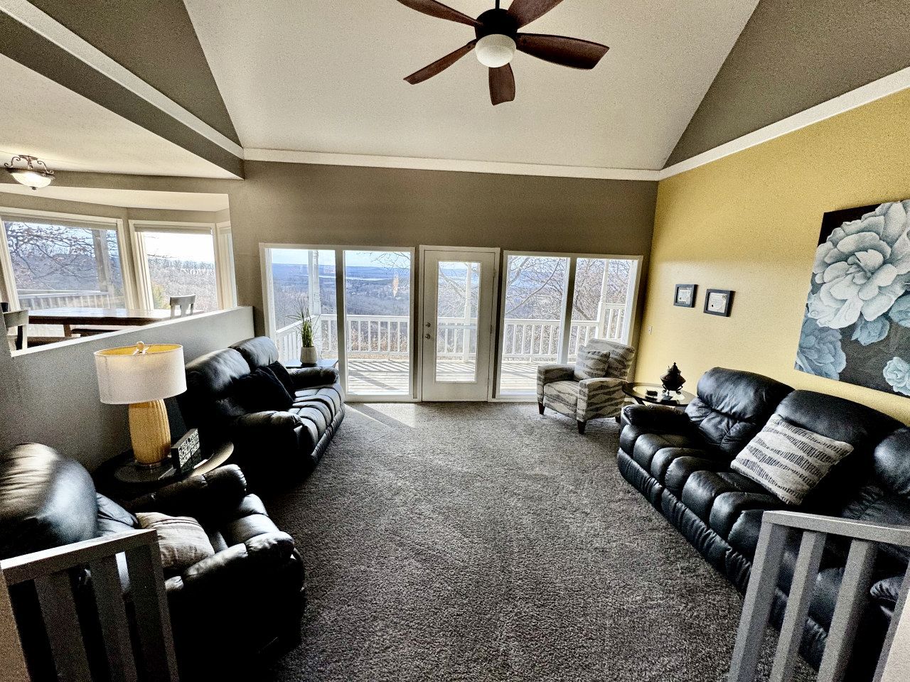 A living room with a ceiling fan, black leather sofas, a lamp, and a large window with a view.