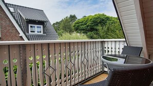 A balcony with a glass table and chairs, a wooden fence with circular cutouts, and a view of a brick building and green trees. - Quietly situated vacation apartment - Ferienhaus Weingarten FeWo zum Fischer - (Baabe)