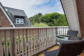 Ein Balkon mit Glastisch und -stühlen, einem Holzzaun mit Rundungen und Blick auf ein Backsteingebäude und grüne Bäume.