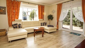 Ein Wohnzimmer mit einem beigefarbenen Ecksofa, einem Holztisch und einem großen Fenster mit orangefarbenen Vorhängen.