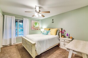 Ein Schlafzimmer mit einem Bett, einem Nachttisch, einem Tisch, einem Deckenventilator und einem Fenster mit Vorhängen.