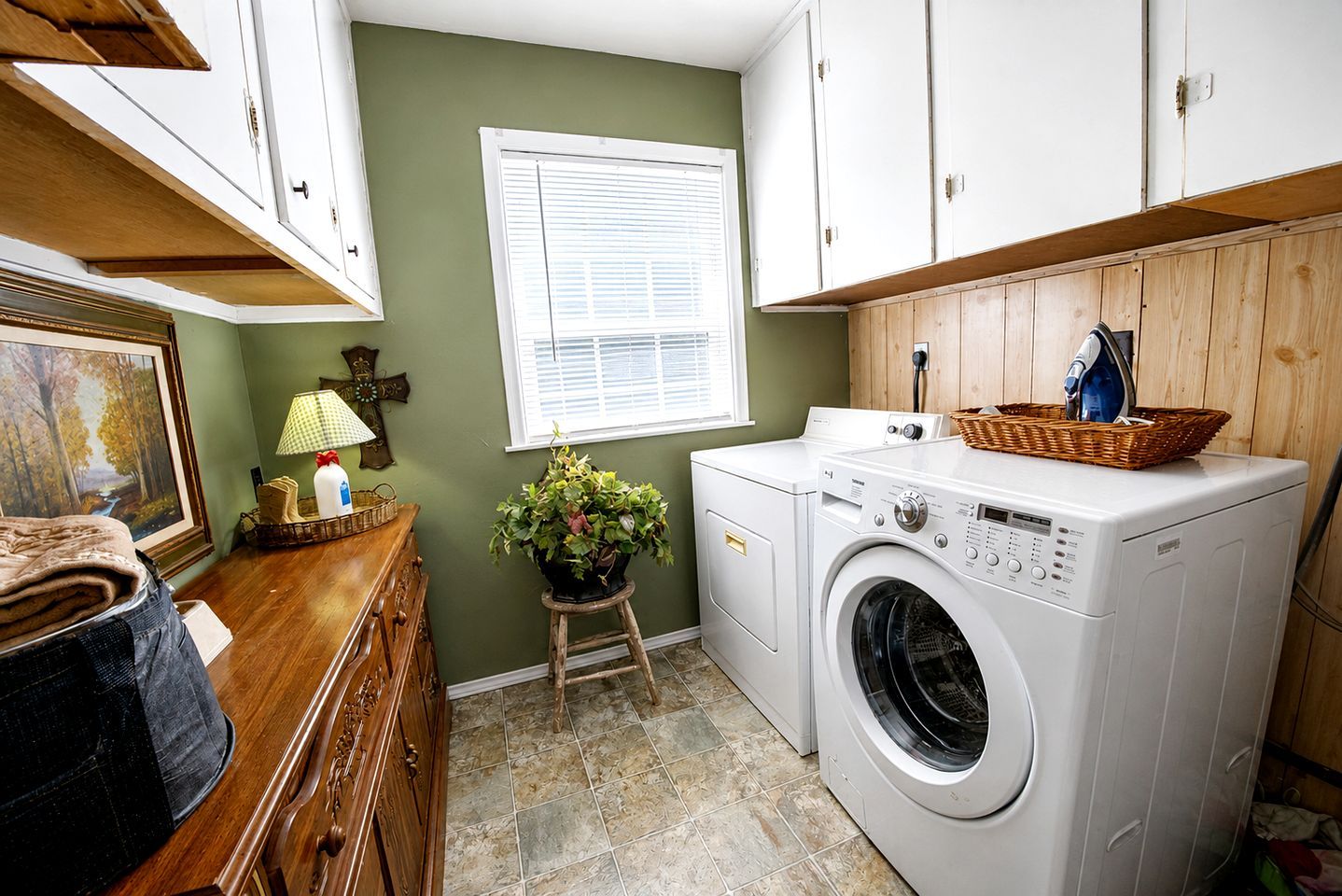 A laundry room with a washing machine, a wooden cabinet, a window with blinds, a small stool with a plant, and a basket on the washing machine.