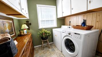 A laundry room with a washing machine, a wooden cabinet, a window with blinds, a small stool with a plant, and a basket on the washing machine.