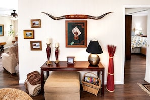 A living room with a wooden table, a lamp, a large horn decoration, framed pictures, and a wicker basket. - Lovely Cottage Rental with Waterfall on San Gabriel River near Austin, Texas (Georgetown)