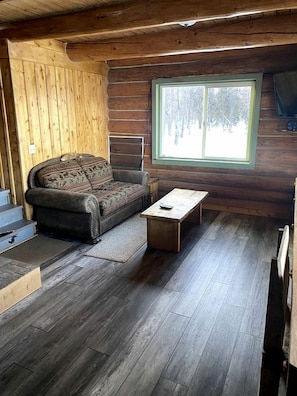 A log cabin interior with a sofa, a small table, and a window with a view of trees. - Off-Grid Cabin with Views from the Porch in Alaska (Cooper Landing)