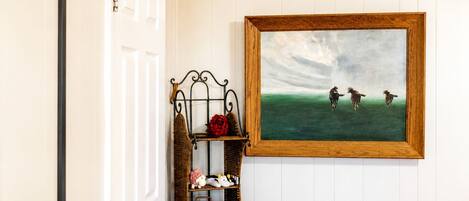 A hallway with a white door, a wooden-framed painting of horses, a wicker bench, and a red rose.