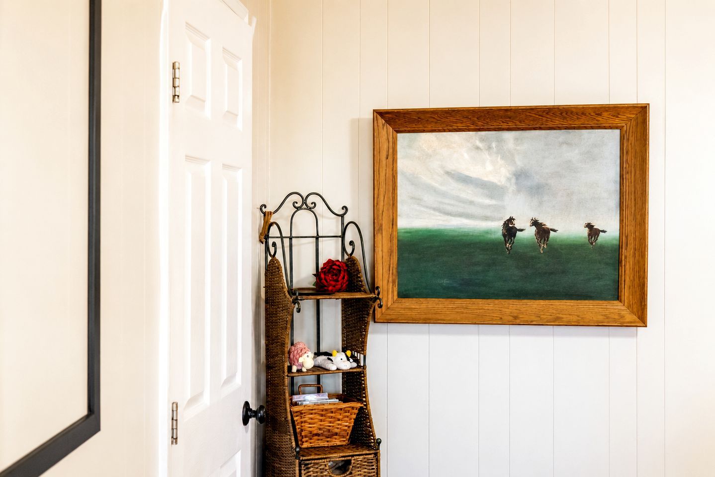 A hallway with a white door, a wooden-framed painting of horses, a wicker bench, and a red rose.