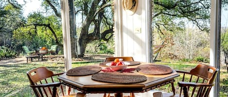 A dining area with a round table, four chairs, and a view of a garden with trees and a bench.