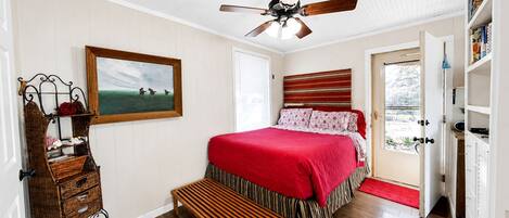 Um quarto com uma cama, um ventilador de teto, uma janela, uma porta e um pequeno banco.