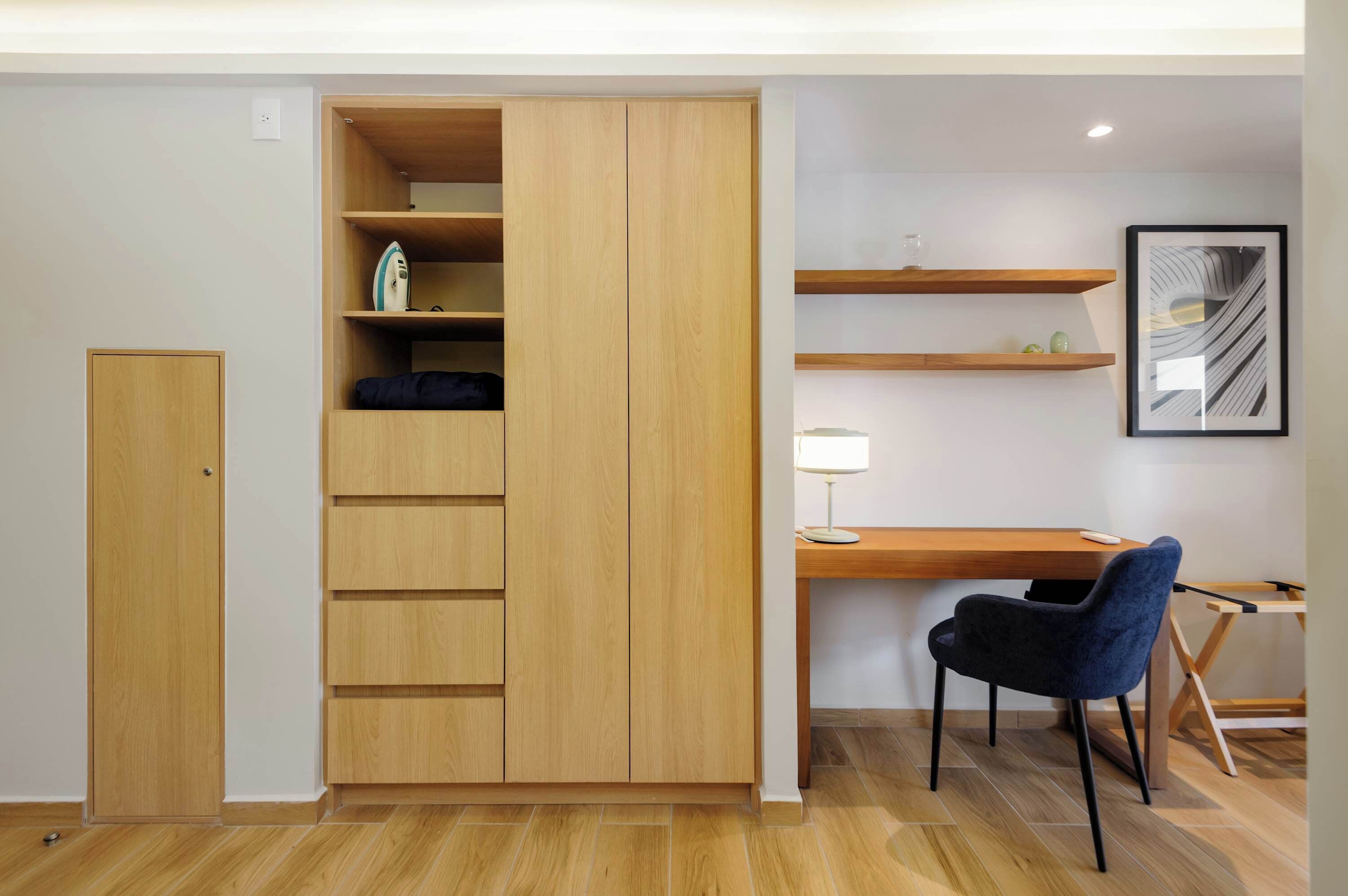 A room with a wooden wardrobe, a desk with a chair, and shelves with decorative items.