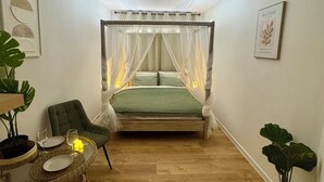 Ein Schlafzimmer mit Baldachinbett, einer kleinen Sitzecke mit Sessel und Tisch sowie zwei Kübelpflanzen.