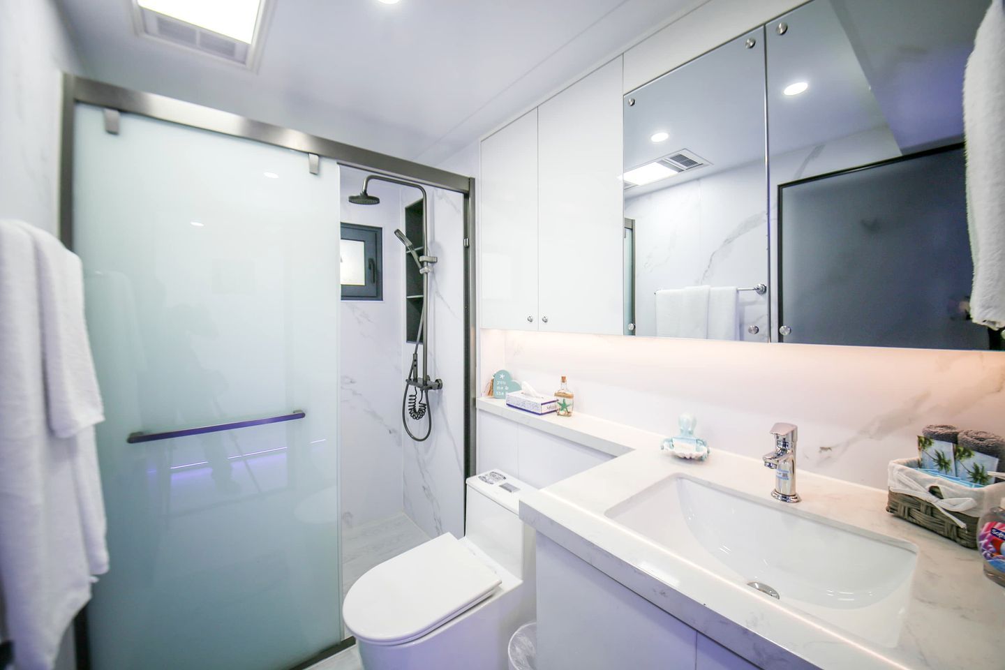 Um banheiro moderno com chuveiro em box de vidro, vaso sanitário branco, pia com bancada de mármore e um espelho grande acima do armário do espelho.