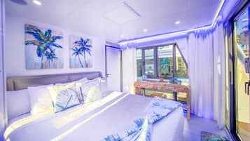 A modern bedroom with a large bed, a small table with decorative items, and a view of the outside through a glass door.
