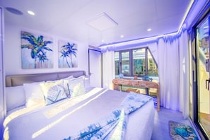 A modern bedroom with a large bed, a small table with decorative items, and a view of the outside through a glass door.
