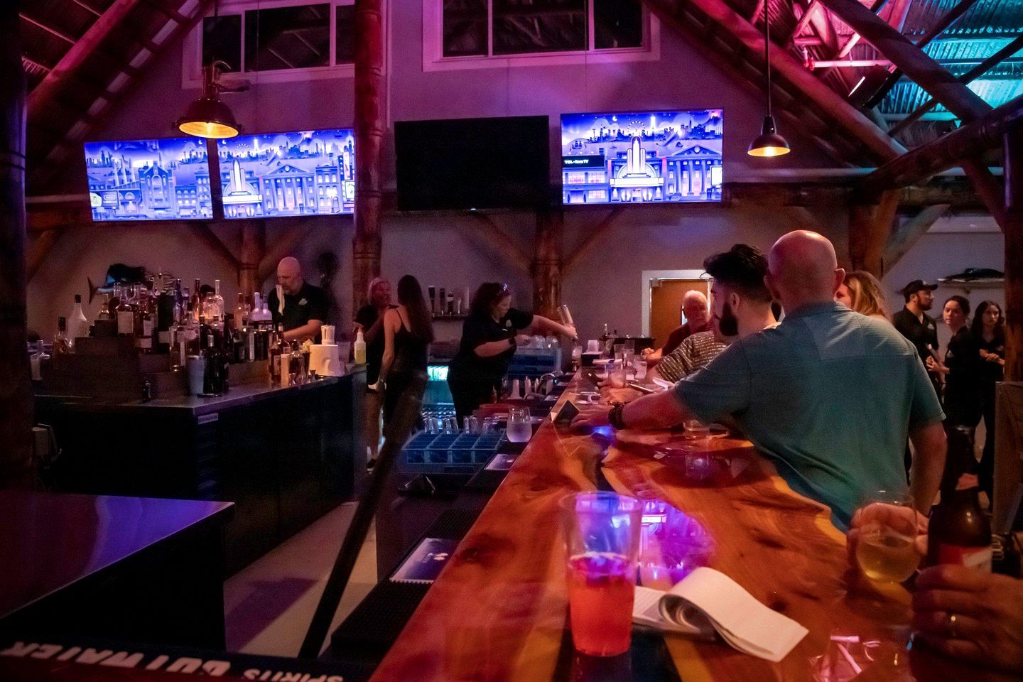 A bar area with multiple screens displaying cityscapes, a bar counter with glasses and bottles, and a group of people socializing.