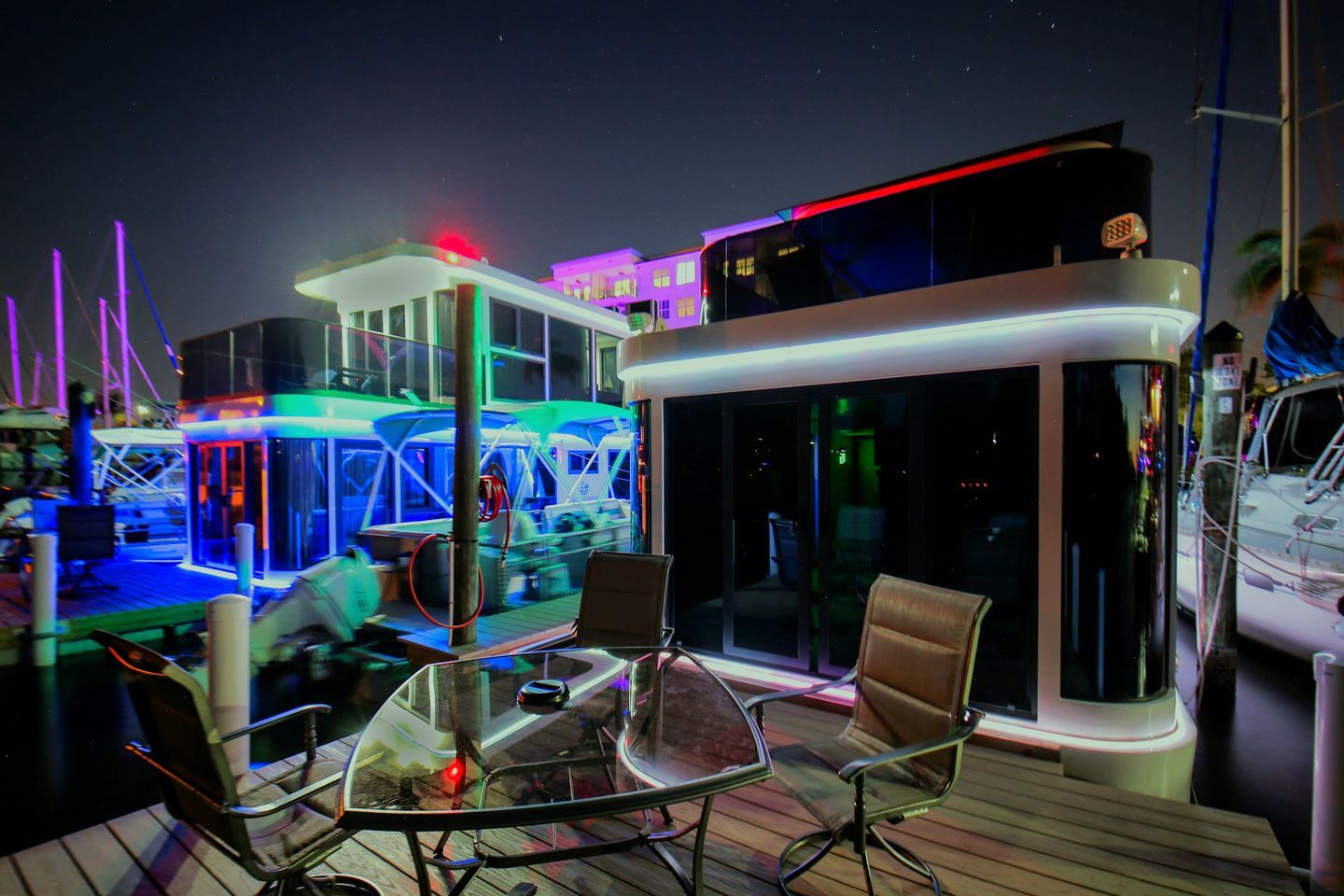 A deck with a glass table and chairs, a control room with monitors, and a building with illuminated windows.