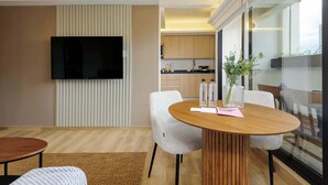 Een moderne woonkamer met een ronde houten tafel, twee witte stoelen, een flatscreen televisie aan de muur en een keuken die zichtbaar is door een glazen wand.