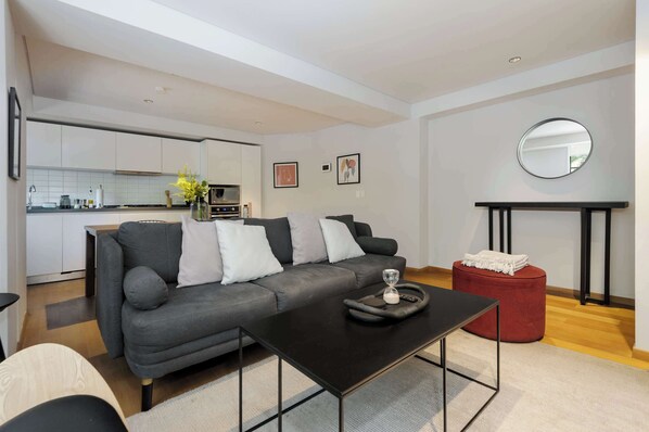 Ein modernes Wohnzimmer mit einem grauen Sofa, einem schwarzen Couchtisch und einem roten Hocker. Im Hintergrund ist eine Küche mit weißen Schränken und einer gefliesten Spritzschutzwand zu sehen.