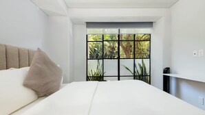 Un dormitorio moderno con una cama grande, una ventana con vista a follaje y un estante pequeño con un dispositivo.