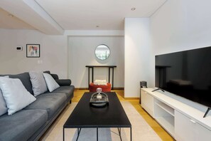 Ein modernes Wohnzimmer mit grauem Sofa, einem schwarzen Couchtisch, einem Fernseher auf einem weißen Schrank und einem roten Hocker.