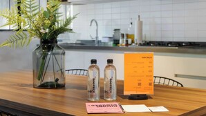 Una mesa de madera con un florero de plantas verdes, dos botellas transparentes con una etiqueta, una tarjeta rosa y un atril para menús.