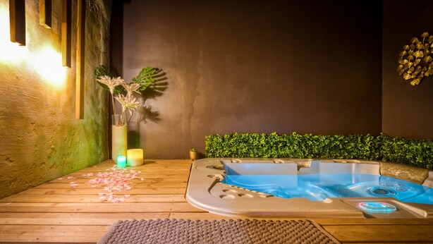 Een wellnessruimte met een jacuzzi, houten vloeren, een decoratieve vaas met bloemen, kaarsen en een groene muur met bladmotief.