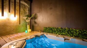 Een hottub met een houten terras, een muur met verticale latten en een groene haag.