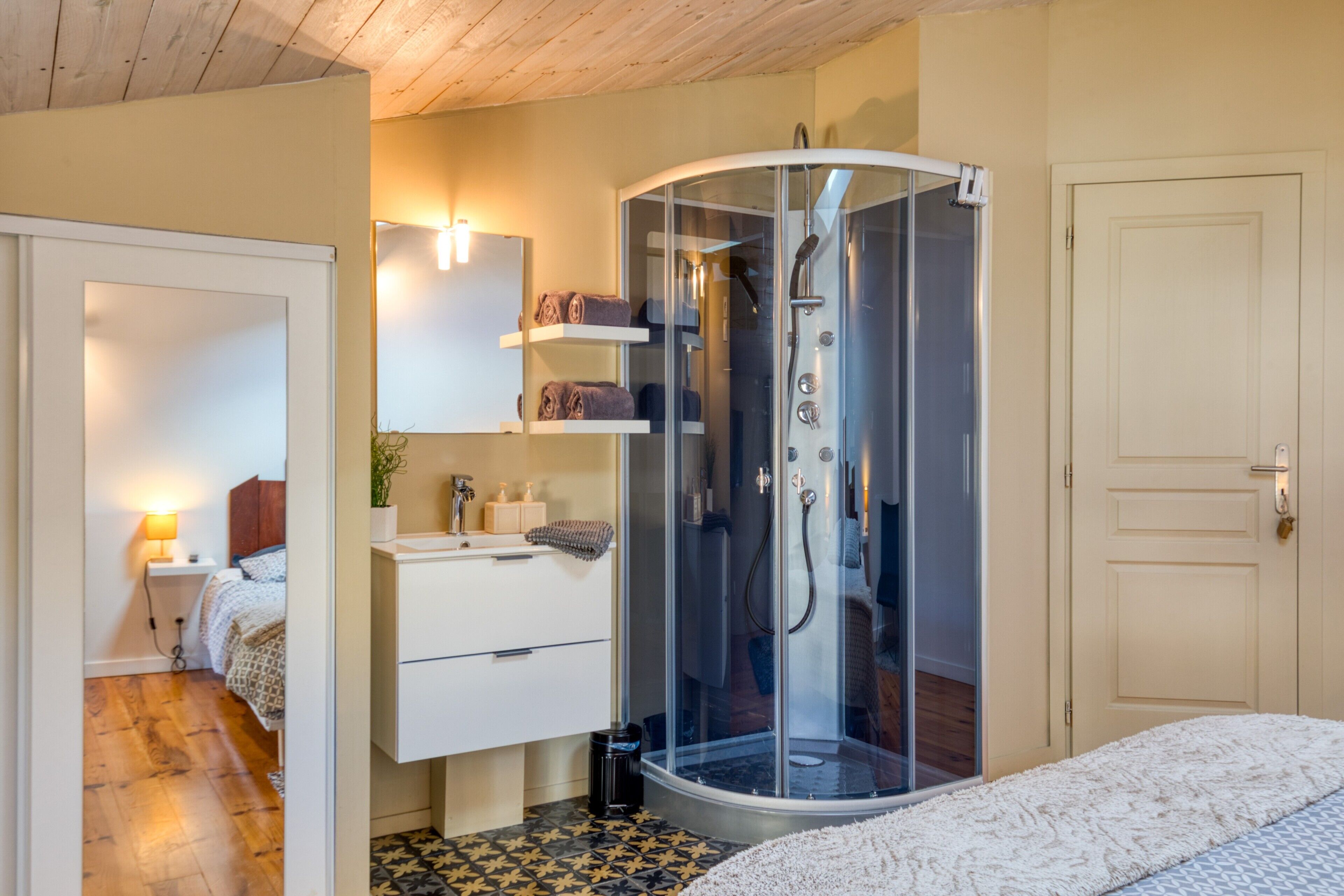 A bedroom with a glass-enclosed shower, a bed with patterned bedding, a bedside table with a lamp, and a door to another room.