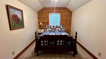 A bedroom with a wooden ceiling fan, a bed with a checkered bedspread, a nightstand, a window with blinds, and a framed picture on the wall.