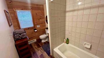A bathroom with a wooden floor, a white bathtub, a toilet, and a window with blinds.
