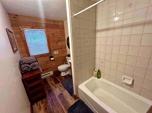 Une salle de bain avec un plancher en bois, une baignoire blanche, des toilettes et une fenêtre avec des stores.